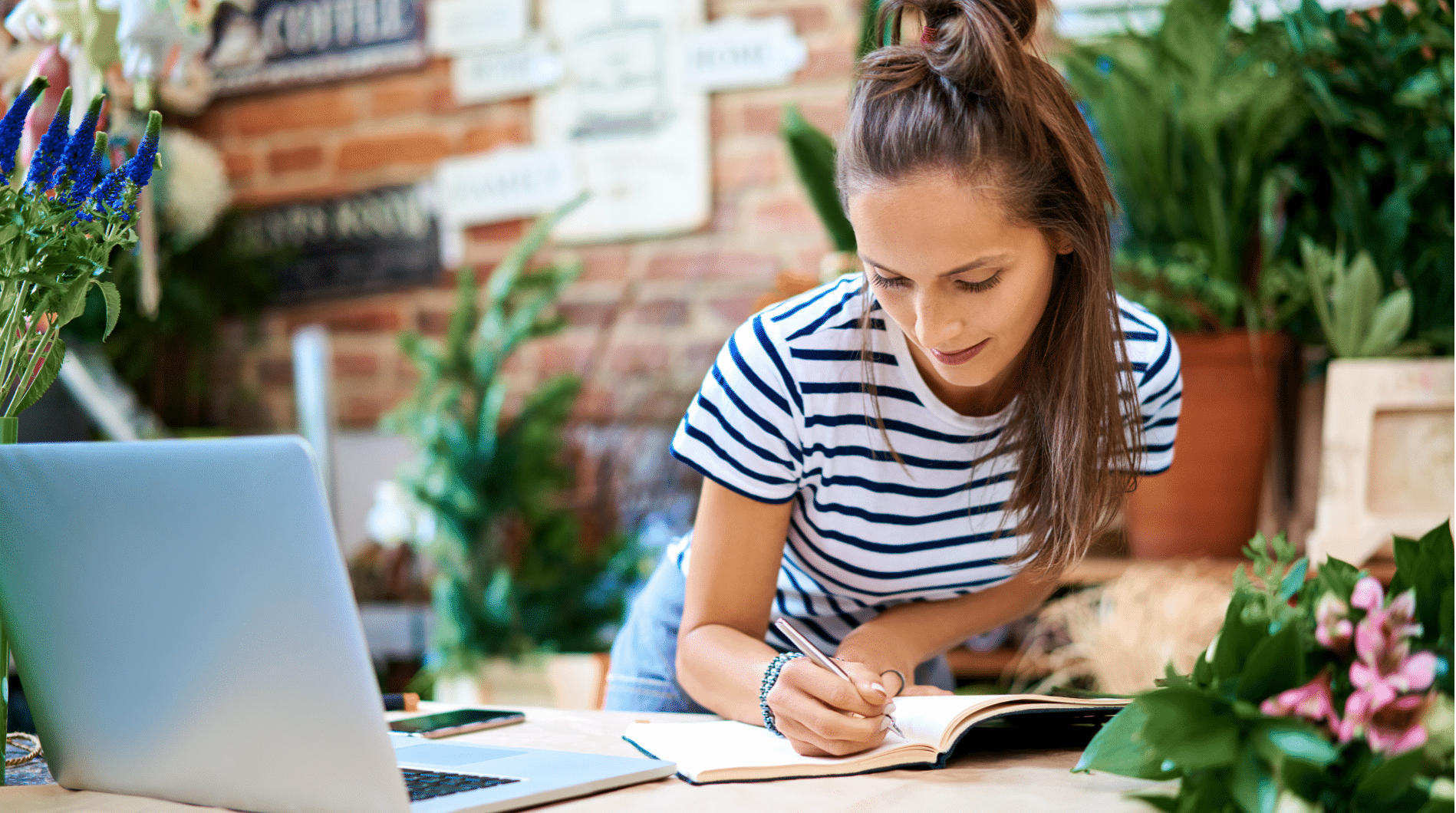 A woman writes in a notebook next to her open laptop.